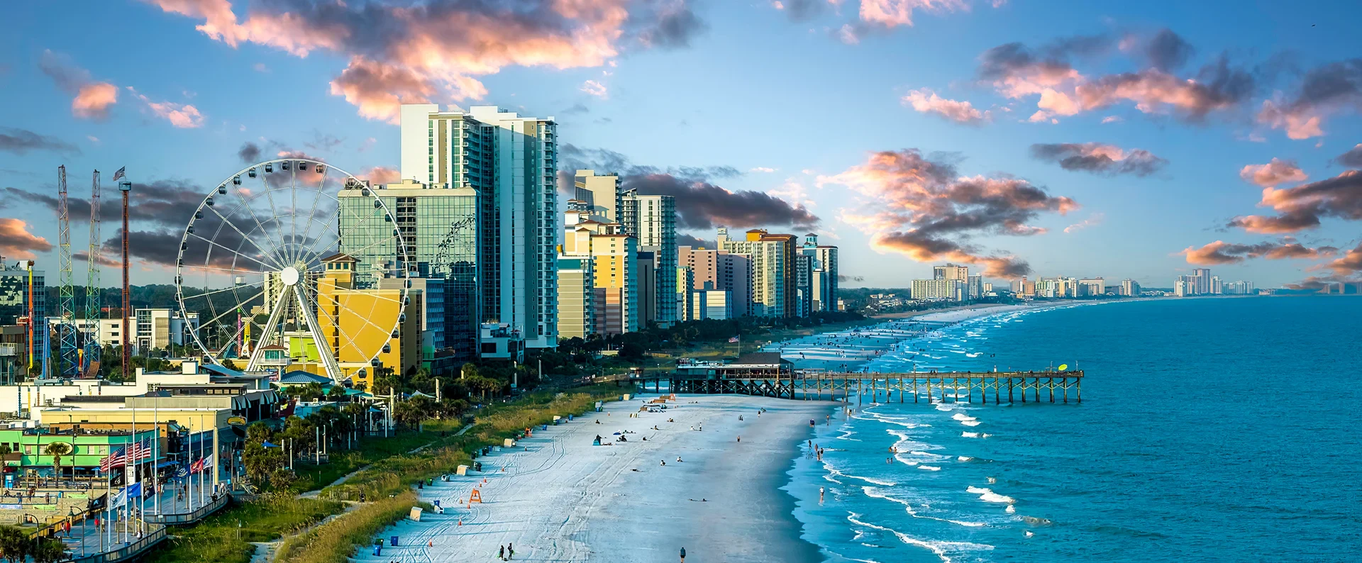 An aerial view of Myrtle Beach, South Carolina, showing the coastline, ocean, pier, and city skyline with buildings and a Ferris wheel.